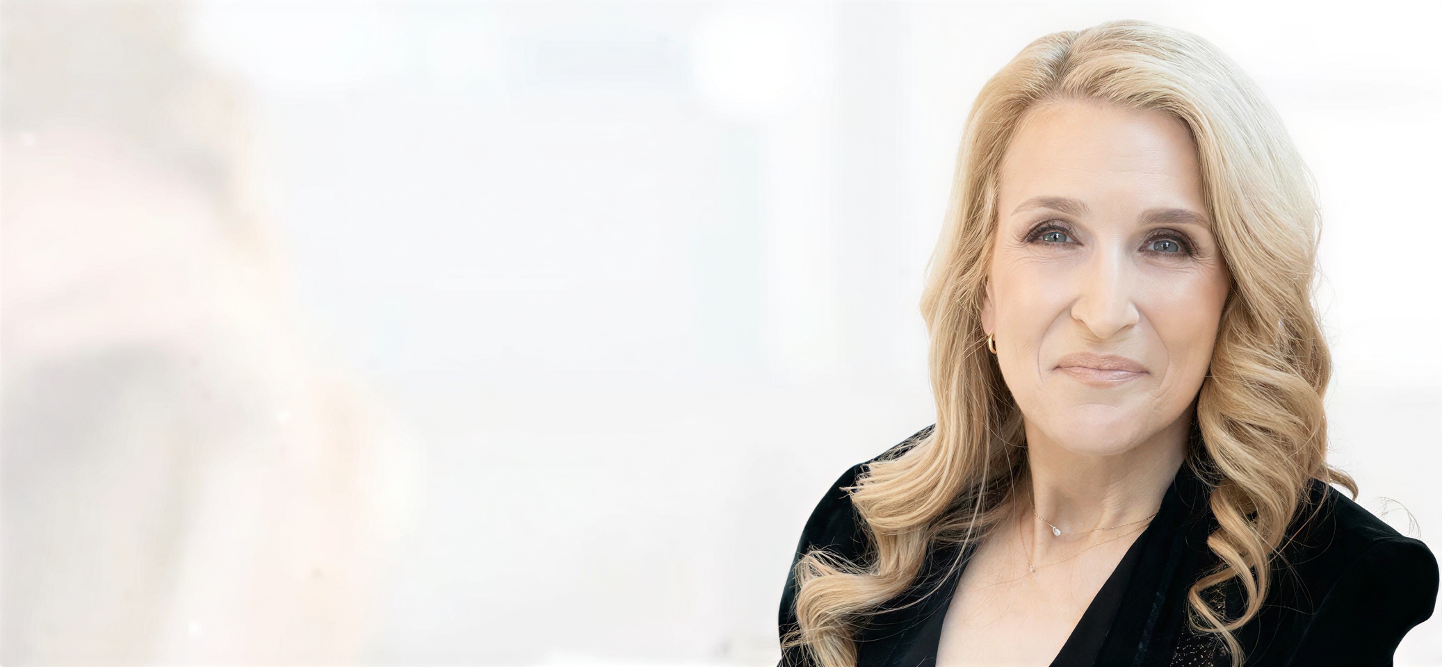 A person with long blonde hair in a black jacket smiles softly.
