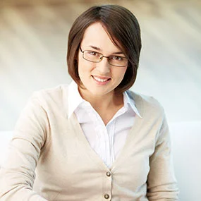 A person with glasses and a beige sweater smiling indoors.