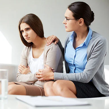 Two women sitting, one comforting the other with a hand on her shoulder.