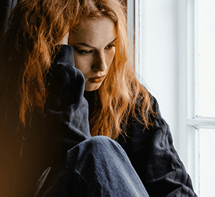 Person with long red hair sitting by a window, looking contemplative.